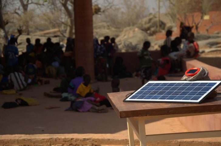 A small solar panel on a table in front of a group of school children under a tree.